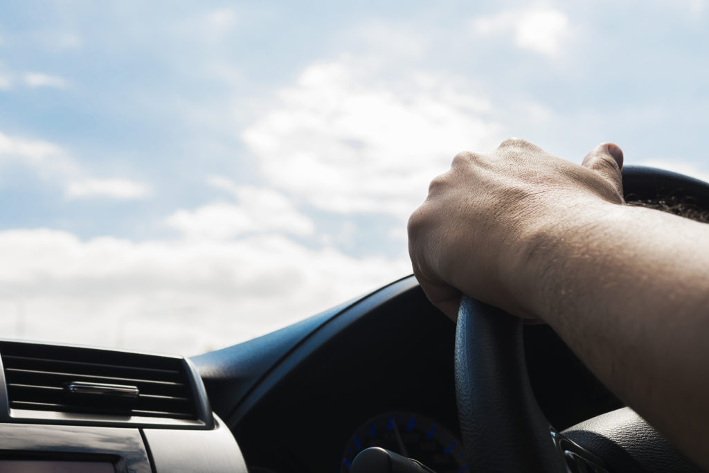 Na imagem há uma pessoa com a mão segurando o volante de um carro a gás, olhando pelo vidro o céu azul com algumas nuvens brancas.