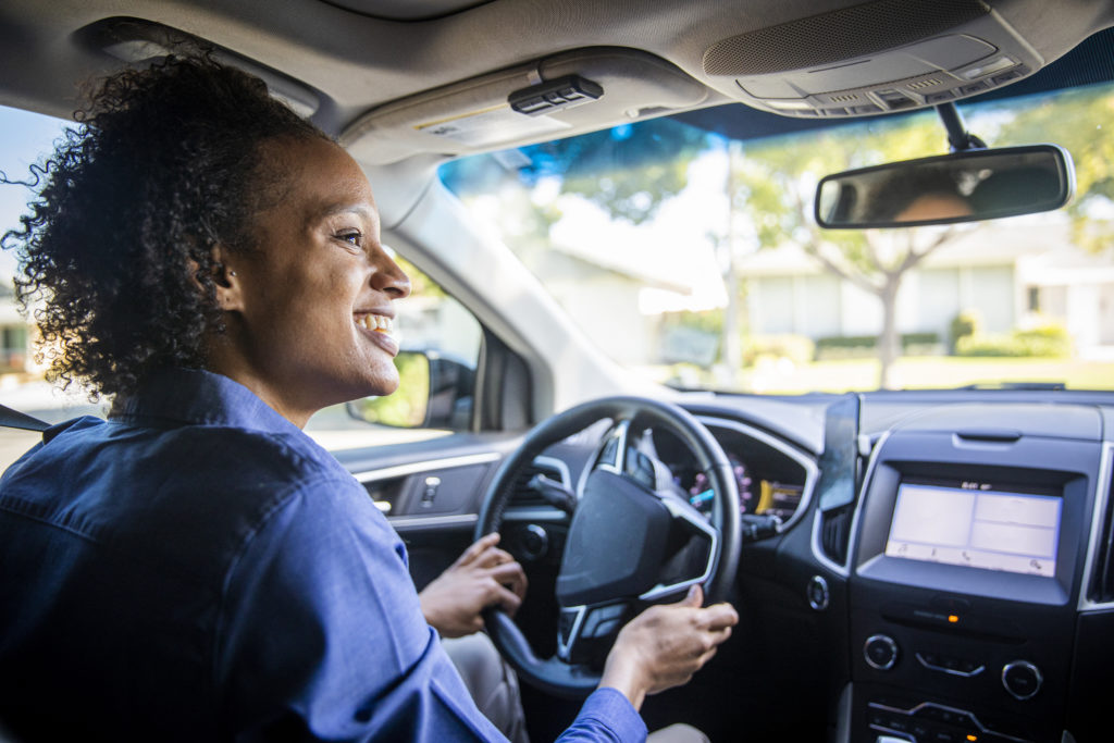 Na imagem há uma mulher negra sorrindo enquanto dirige.