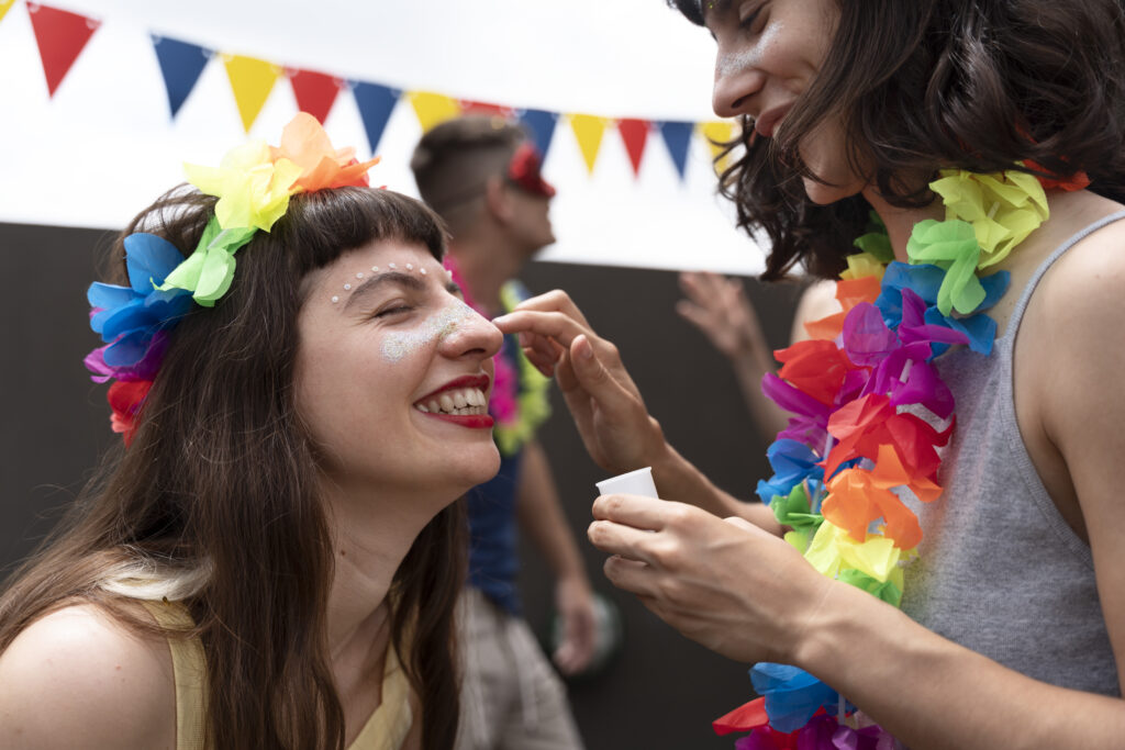 Na imagem há duas pessoas do gênero feminino com itens temáticas de carnaval. Uma delas está maquiando com glitter a outra.
