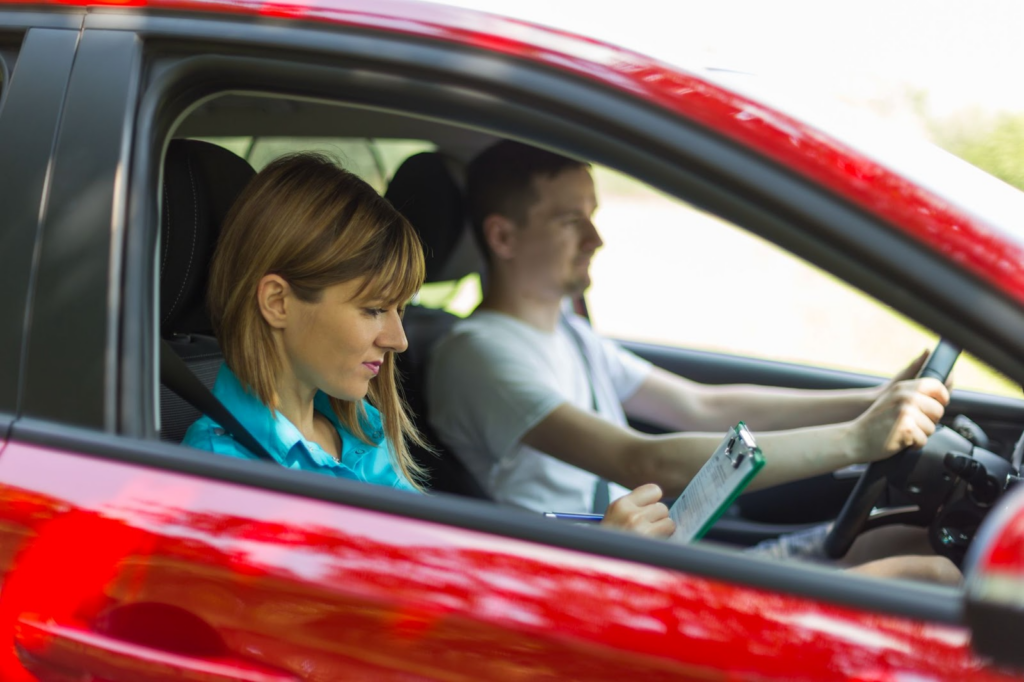 Um carro vermelho com duas pessoas dentro. Ao lado do motorista, uma mulher está segurando uma prancheta realizando anotações