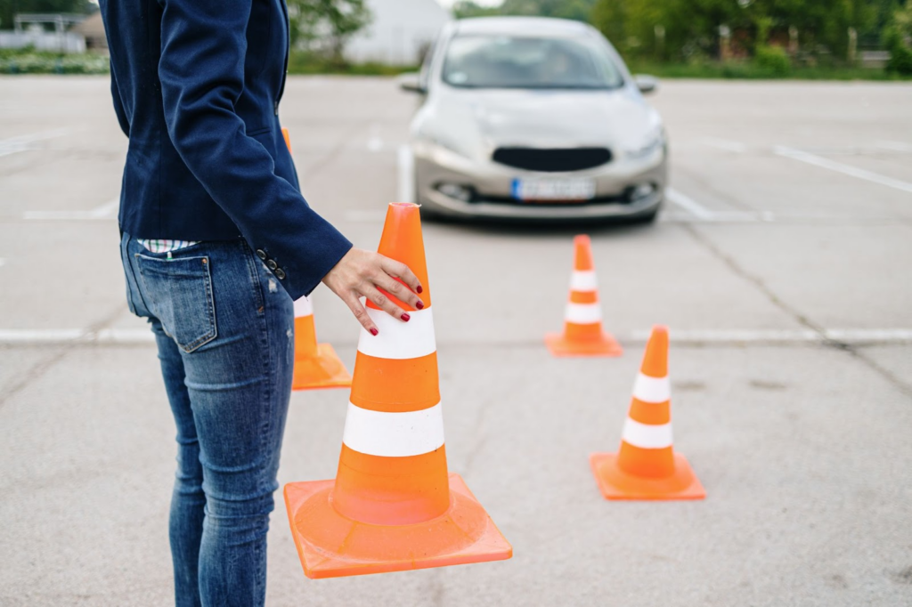 Em um estacionamento, uma pessoa está segurando um cone de trânsito. Outros 3 cones são mostrados na imagem. Ao fundo, um veículo está parado. 