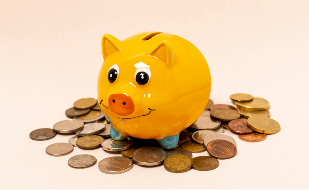 Imagem de um pequeno cofre em formato de porquinho, de cor amarela, patas azuis e nariz laranja. Há moedas espalhadas ao redor do cofre.